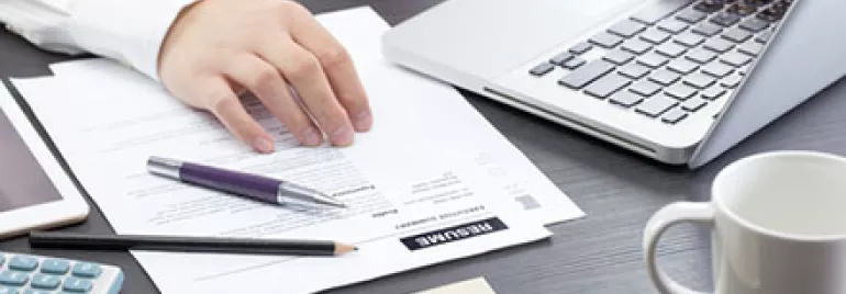 Person working on a laptop at a desk with documents, glasses, a calculator, and a coffee cup.